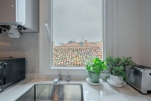 Una cocina con un lavamanos de acero inoxidable, un microondas y un tostador. Se ve una ventana con vista a un tejado de baldosas.