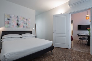 A bedroom with a bed, a white door, a wall-mounted TV, and a dining area with chairs and a table.