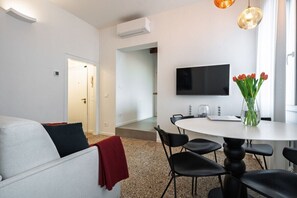 A modern living room with a sofa, a dining table with chairs, a flat-screen TV mounted on the wall, and a vase with flowers.