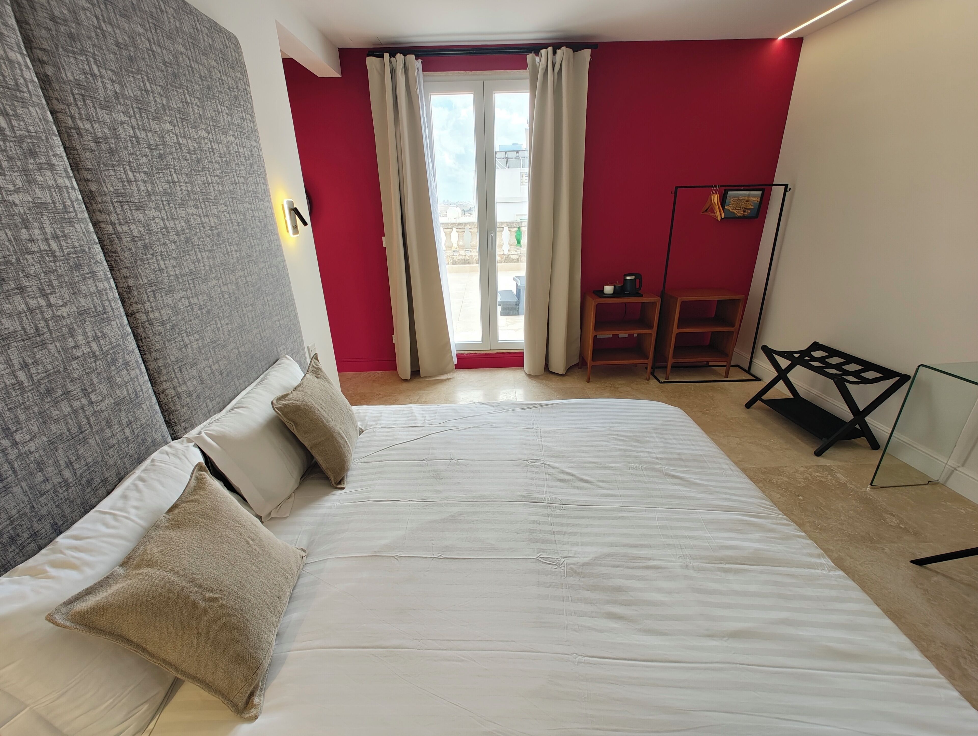 A bedroom with a large bed, a red wall, a window with curtains, and a small table with a cup.