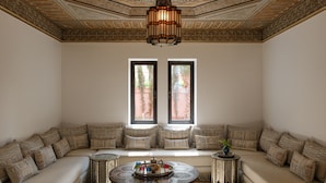 A room with a large, cushioned seating area, a round table with a tray of items, and two windows with a view of greenery. - YALI, private house in the heart of the Four Seasons Resort Marrakech (Marrakech)
