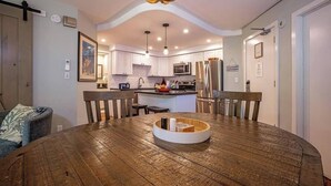 Un comedor con mesa y sillas de madera, una cocina al fondo y una lámpara moderna.