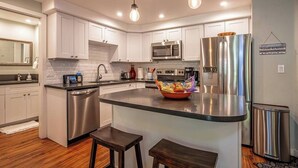 Una cocina moderna con armarios blancos, electrodomésticos de acero inoxidable, una isla central con taburetes y un cartel en la pared.