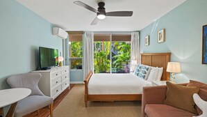 Habitación de hotel con cama, televisión, escritorio, silla, sofá y balcón con vistas a zonas verdes.