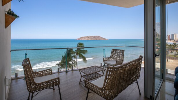 A balcony with wicker furniture, a glass railing, and a view of the ocean and a distant island. - 🌴 Luxury Beachfront Apartment in Mazatlan's Golden Zone - Pool & Oceanfront (Mazatlán)