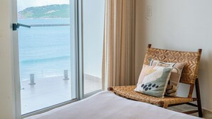 A room with a large window overlooking a body of water, a wicker chair with a cushion, and a plain wall. - 🌴 Luxury Beachfront Apartment in Mazatlan's Golden Zone - Pool & Oceanfront (Mazatlán)