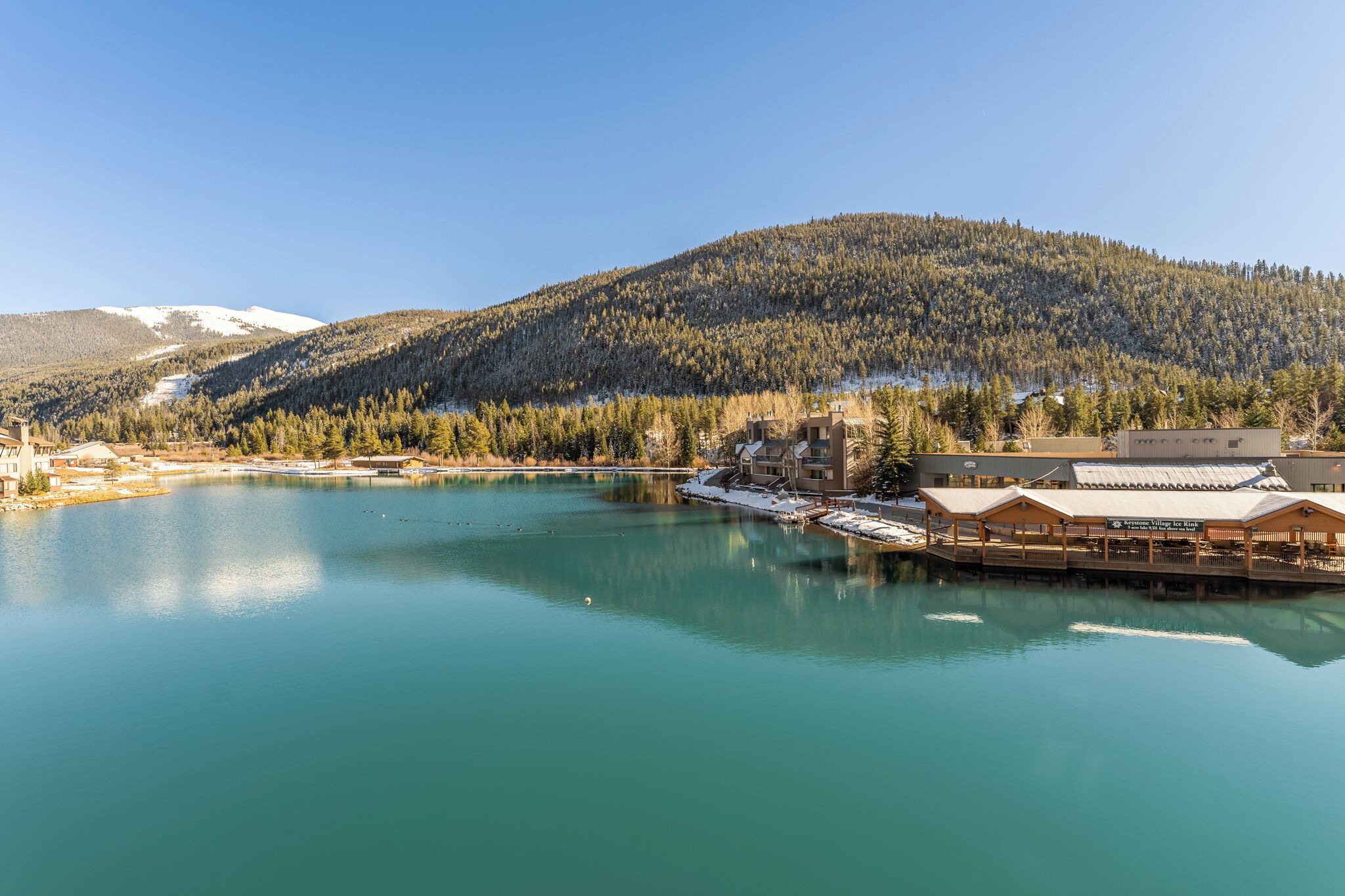 A lakeside hotel with a dock, surrounded by forested hills and a clear sky.