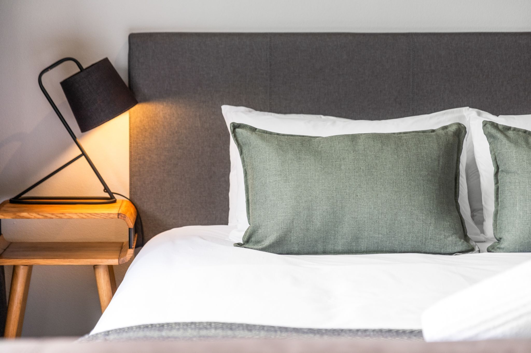 A neatly made bed with white linens and two green pillows, a bedside table with a lamp, and a grey upholstered headboard.