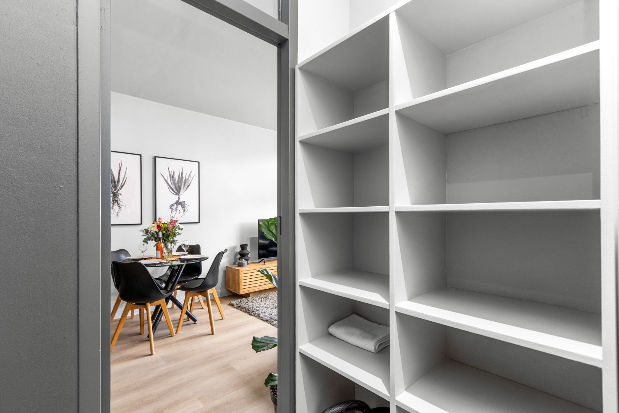 A modern interior with a dining area featuring a glass table and black chairs, a white shelving unit, and framed artwork on the wall.