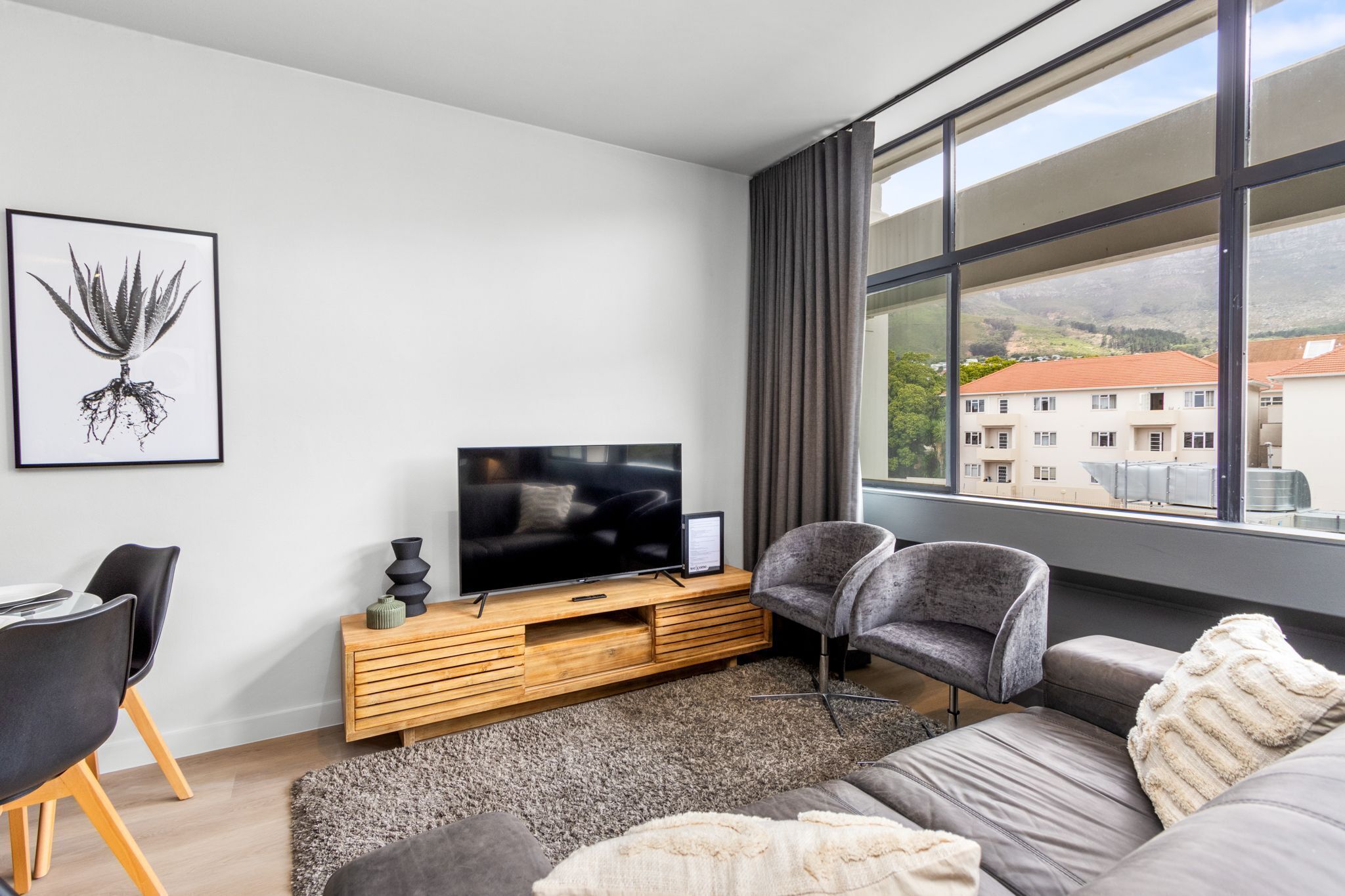A modern living room with a flat-screen TV on a wooden console, a grey sofa, a dining area with a table and chairs, and a large window offering a view of buildings and greenery.