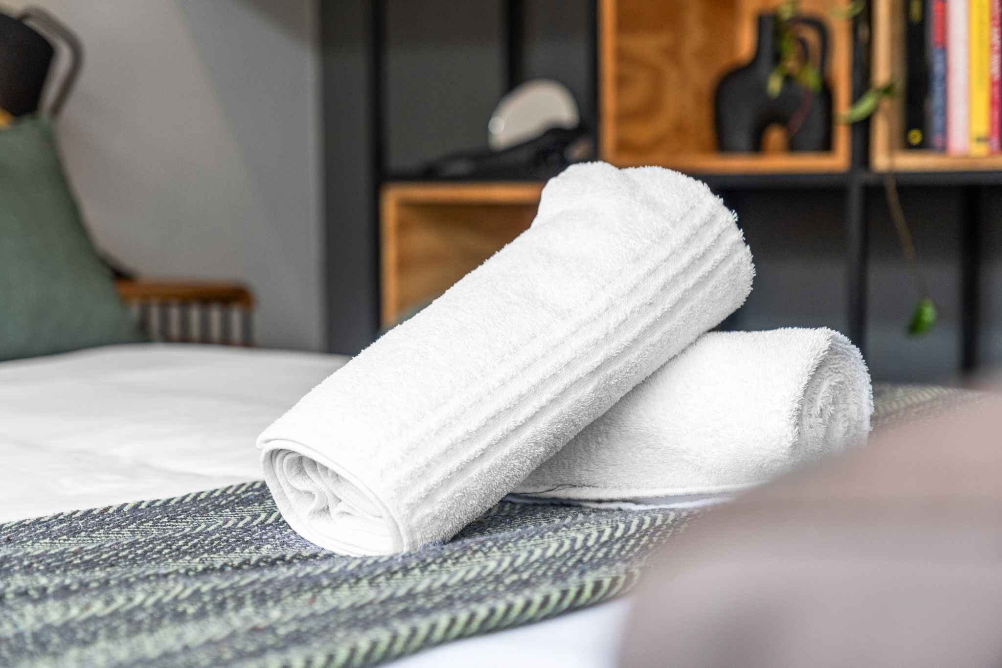 A neatly made bed with a rolled white towel on top, a patterned rug, and a bookshelf in the background.
