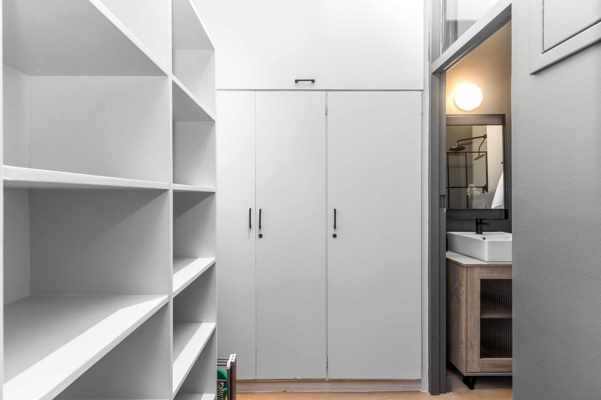 A modern, minimalist walk-in closet with white shelving and doors, a wooden cabinet, and a mirror with a light above it.