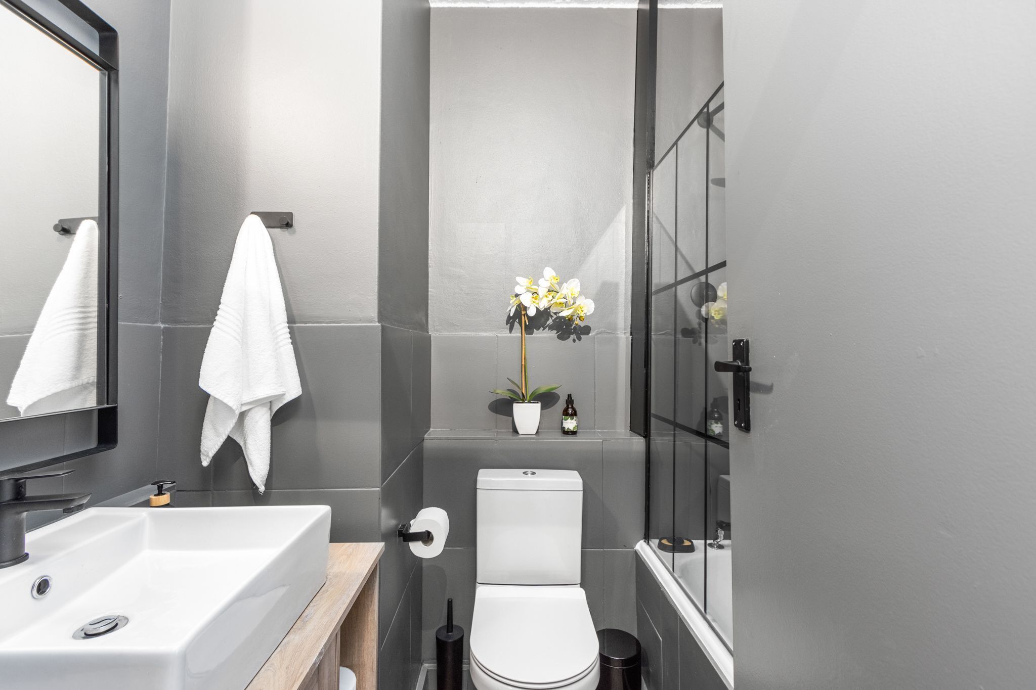 A modern bathroom with a white sink, toilet, and shower. There is a mirror, a towel rack, and a potted plant on the wall.
