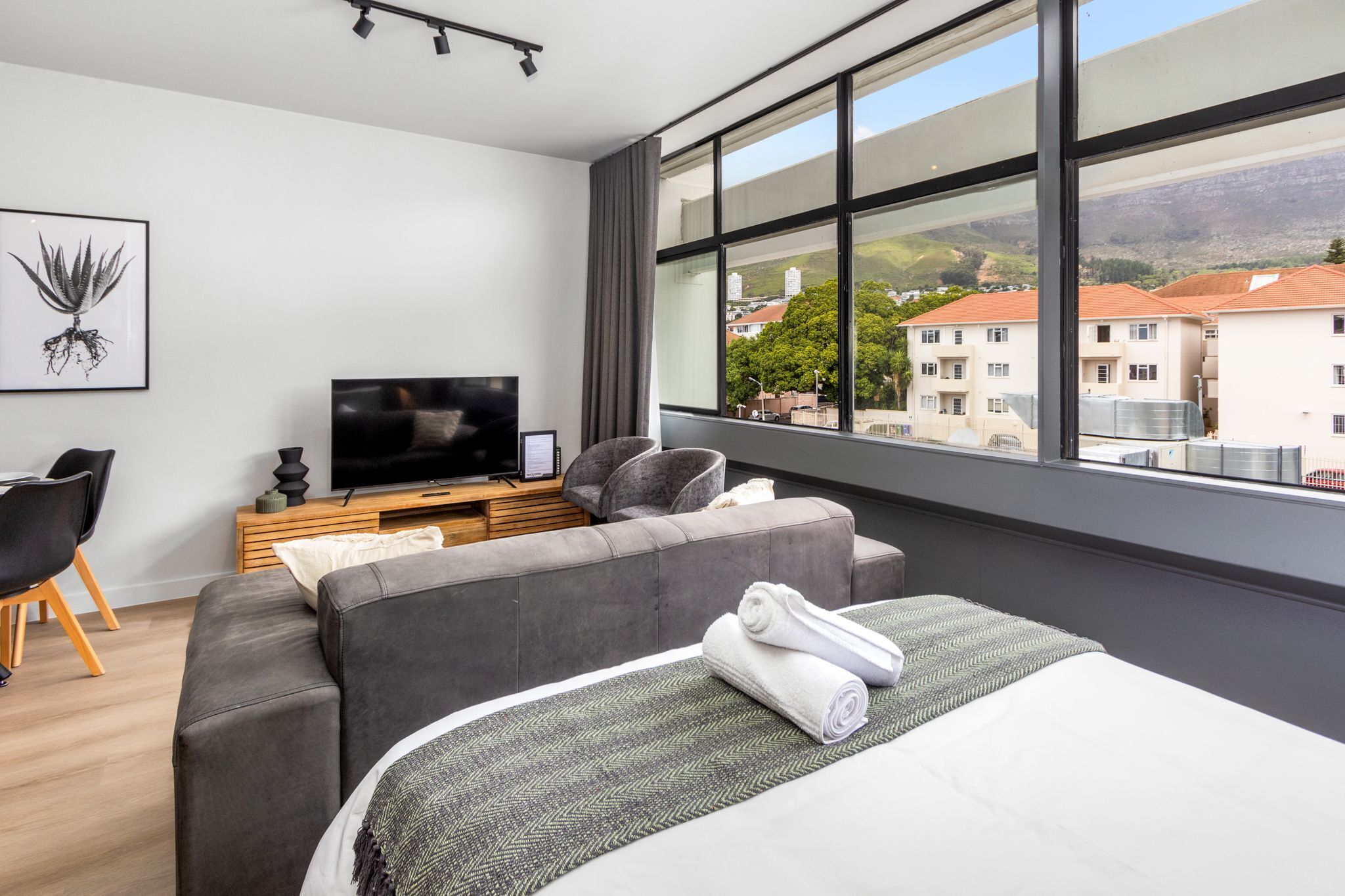 A modern hotel room with a large window, a grey sofa, a flat-screen TV, and a neatly made bed.