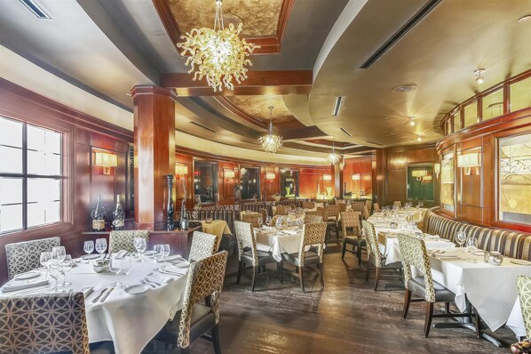 Ein geräumiges Restaurant mit hohen Decken, Kristalllüstern und einer warmen Holzvertäfelung. Die Tische sind mit weißen Tischdecken und Stühlen gedeckt.
