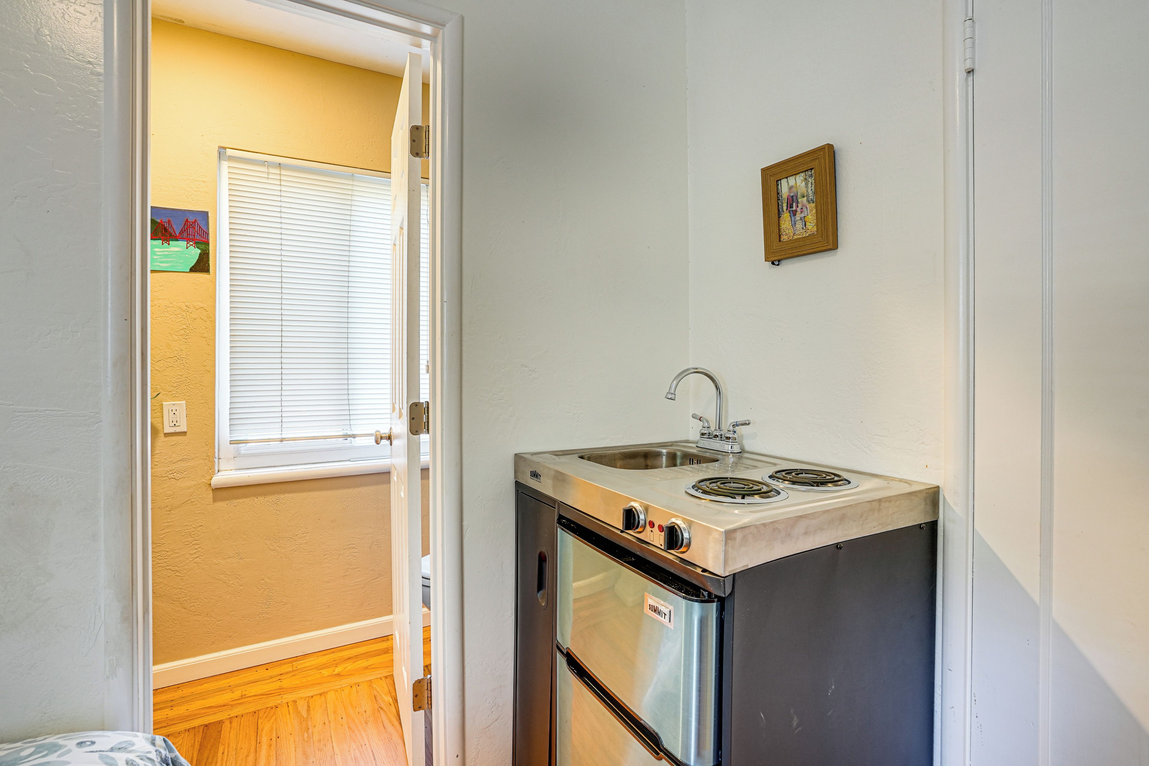 A compact kitchen with a built-in oven and sink, a window with blinds, and a small painting on the wall.