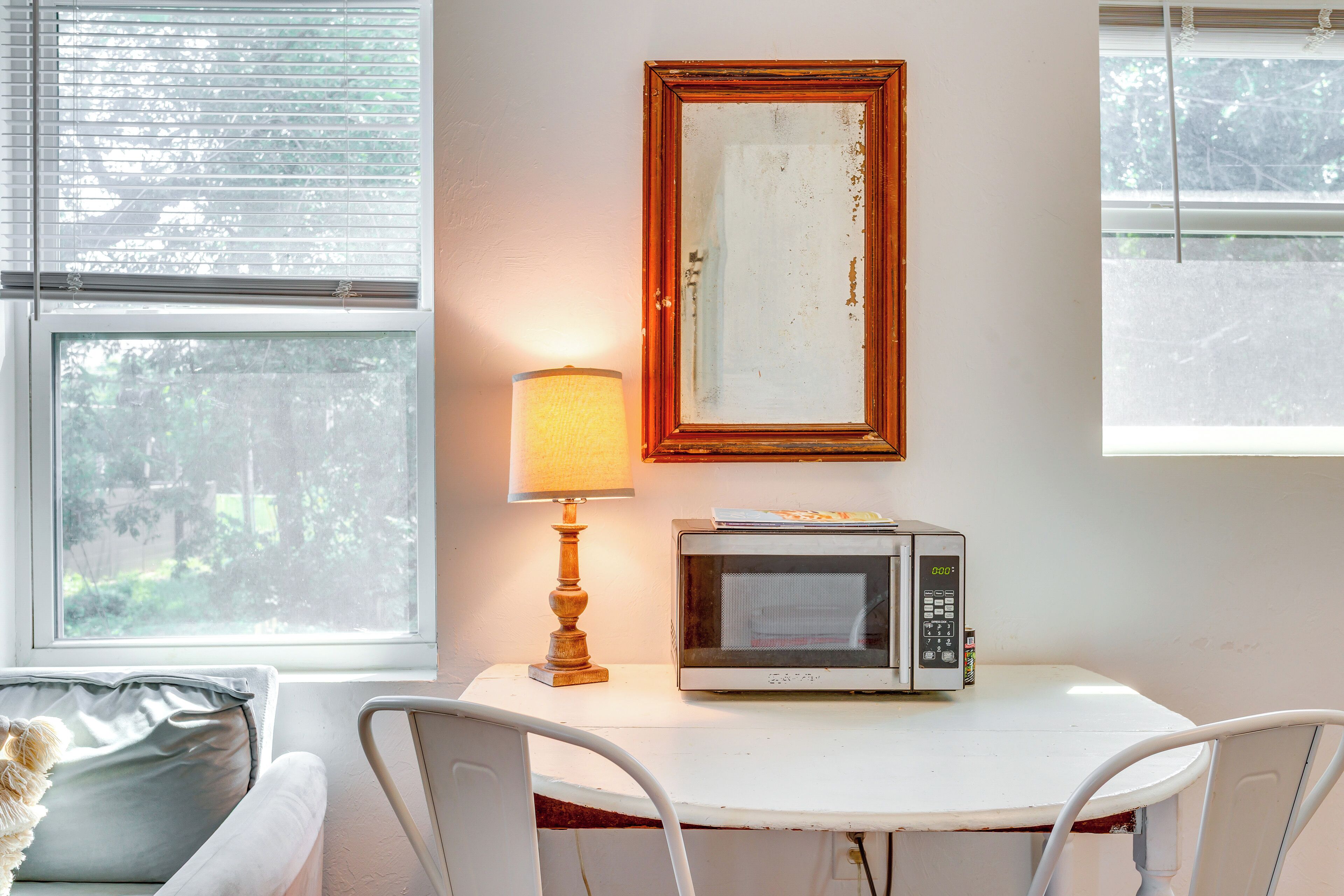 A small dining area with a table, chairs, a lamp, a microwave, and a framed mirror on the wall.