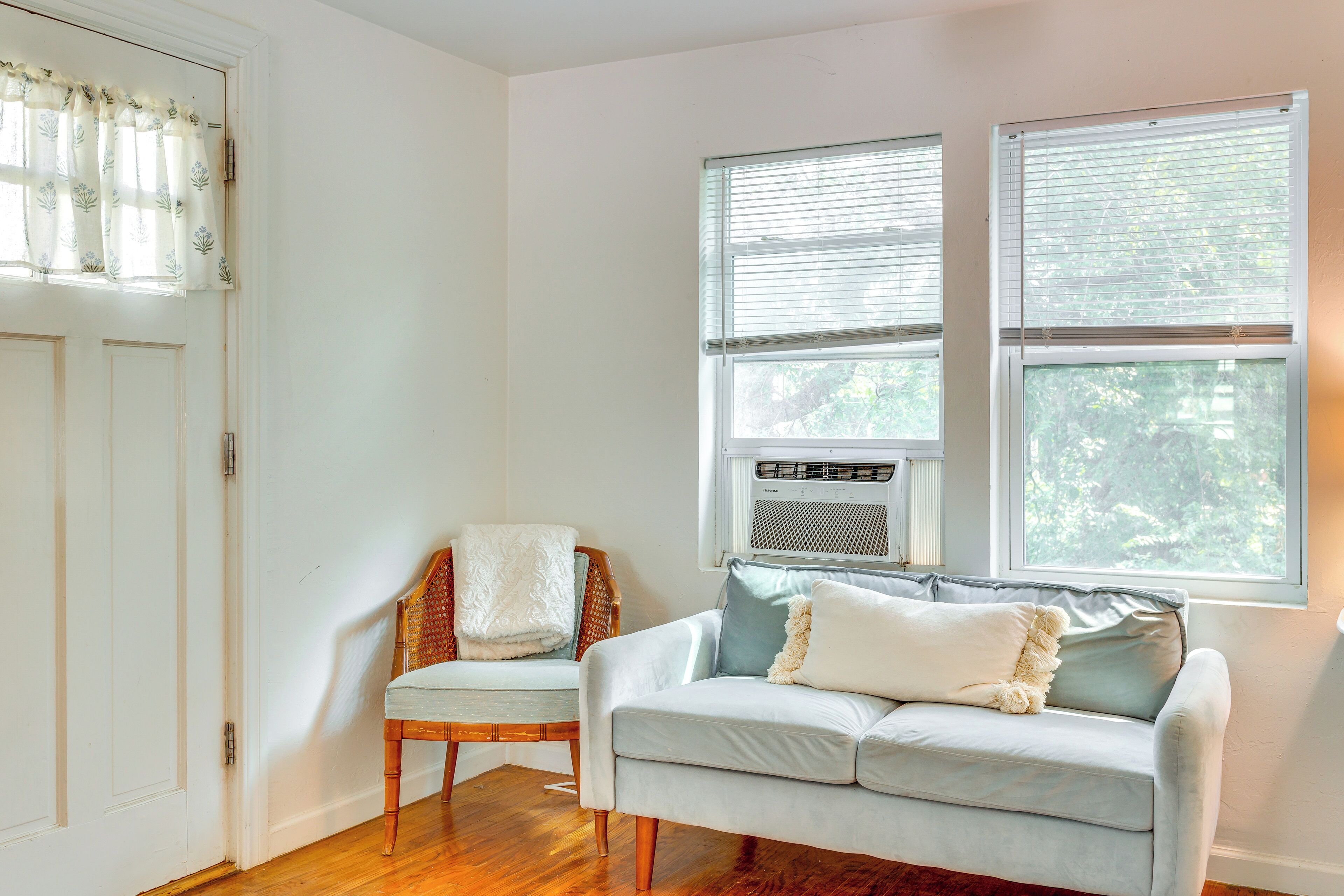 A room with a light blue sofa, a wicker chair, and a window with blinds.