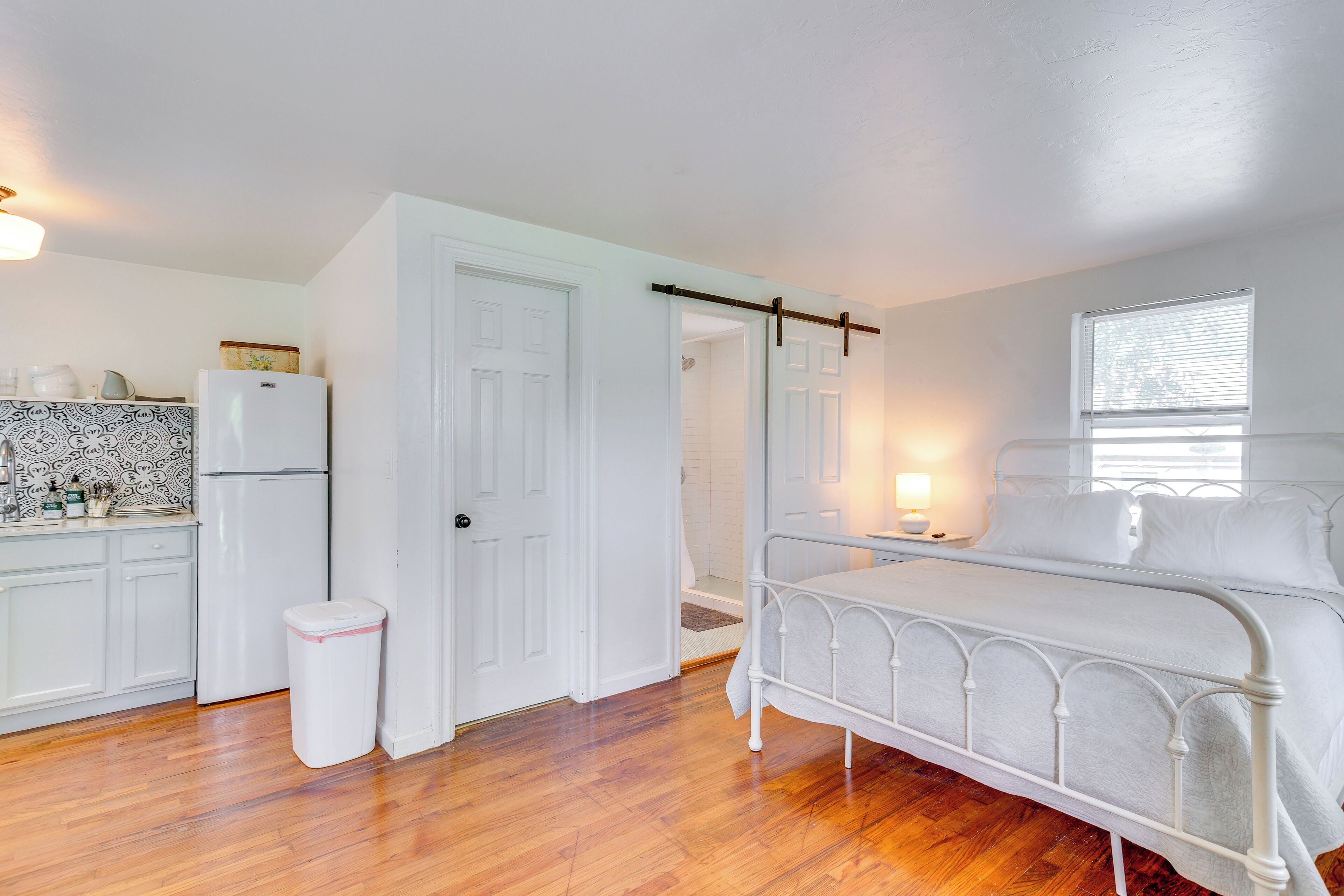A bedroom with a metal bed frame, a white door, a kitchen area with a refrigerator, and a trash bin.