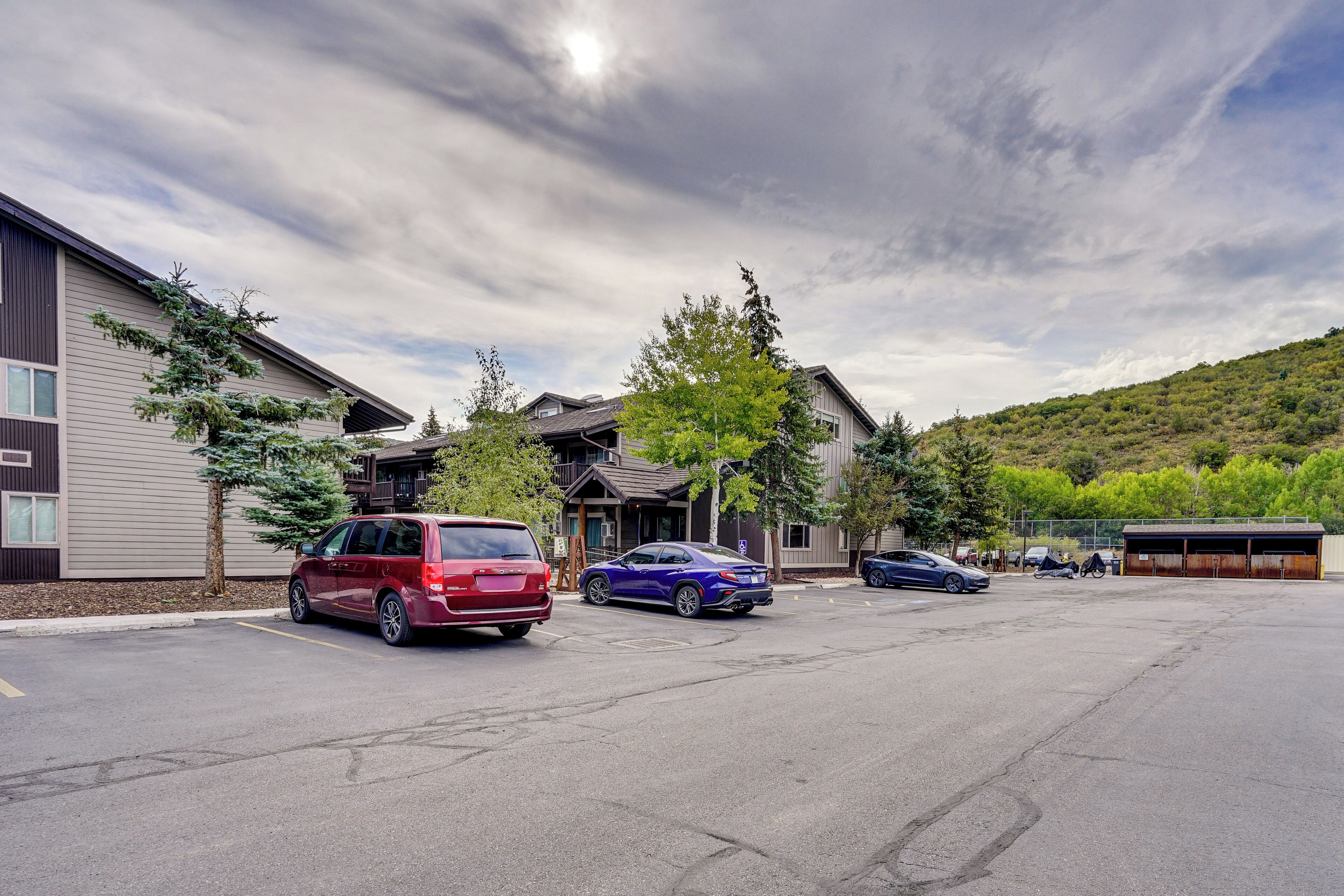 A parking lot with several cars, a building with a sloped roof, and a hilly landscape in the background.
