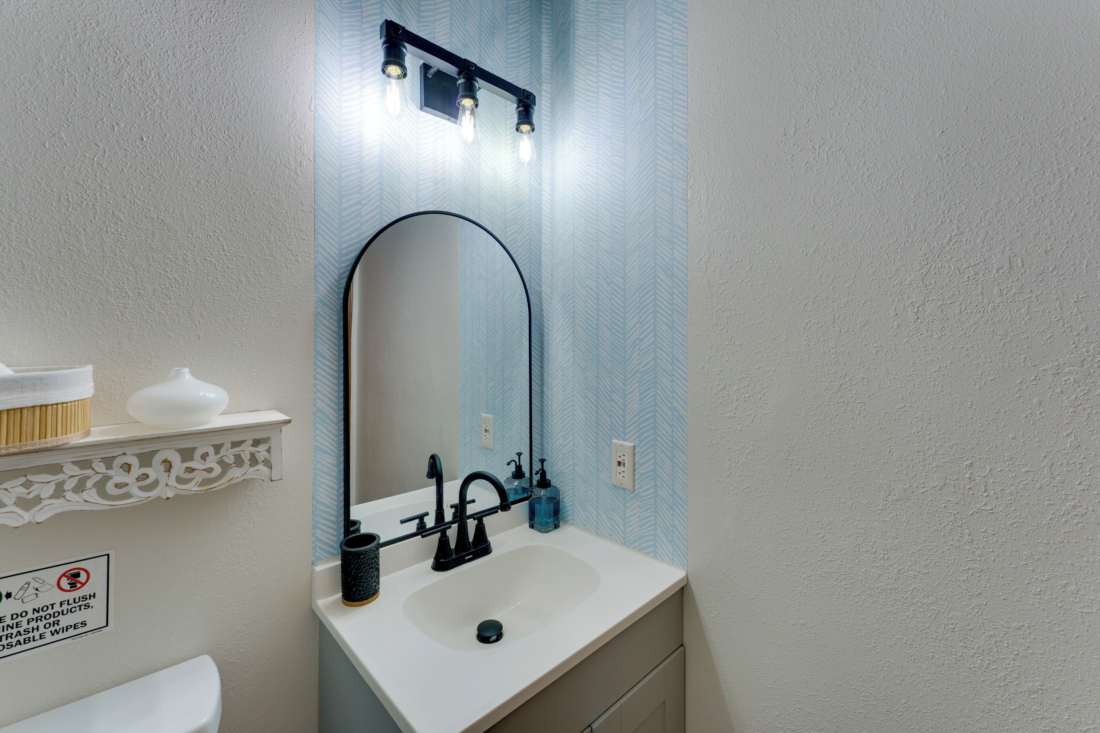 A bathroom with a white sink, black faucet, and a mirror with a curved frame. There is a shelf with a white vase and a sign on the wall.