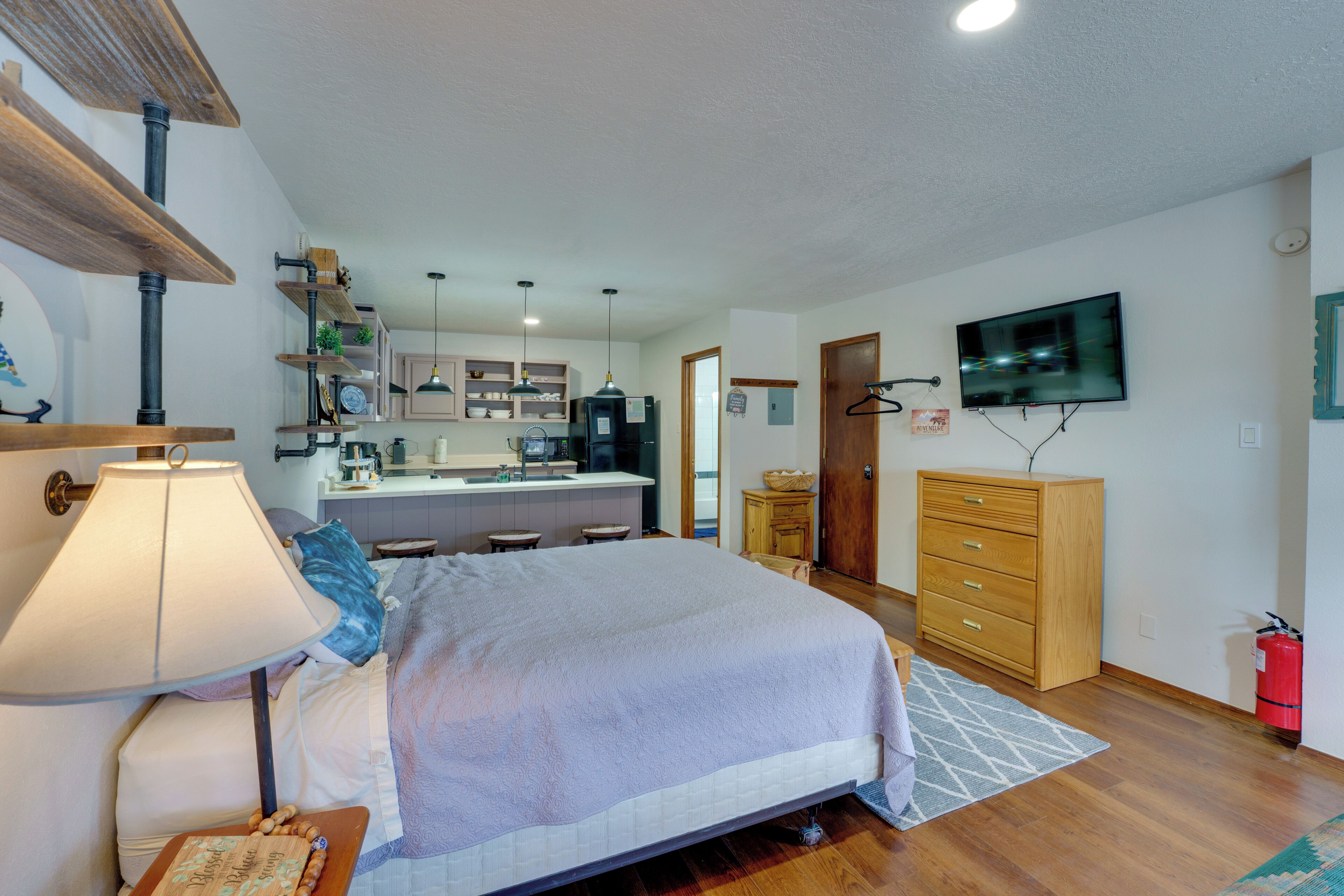 Ein Schlafzimmer mit Bett, Nachttisch, Lampe und Blick auf die Küche und das Wohnzimmer.