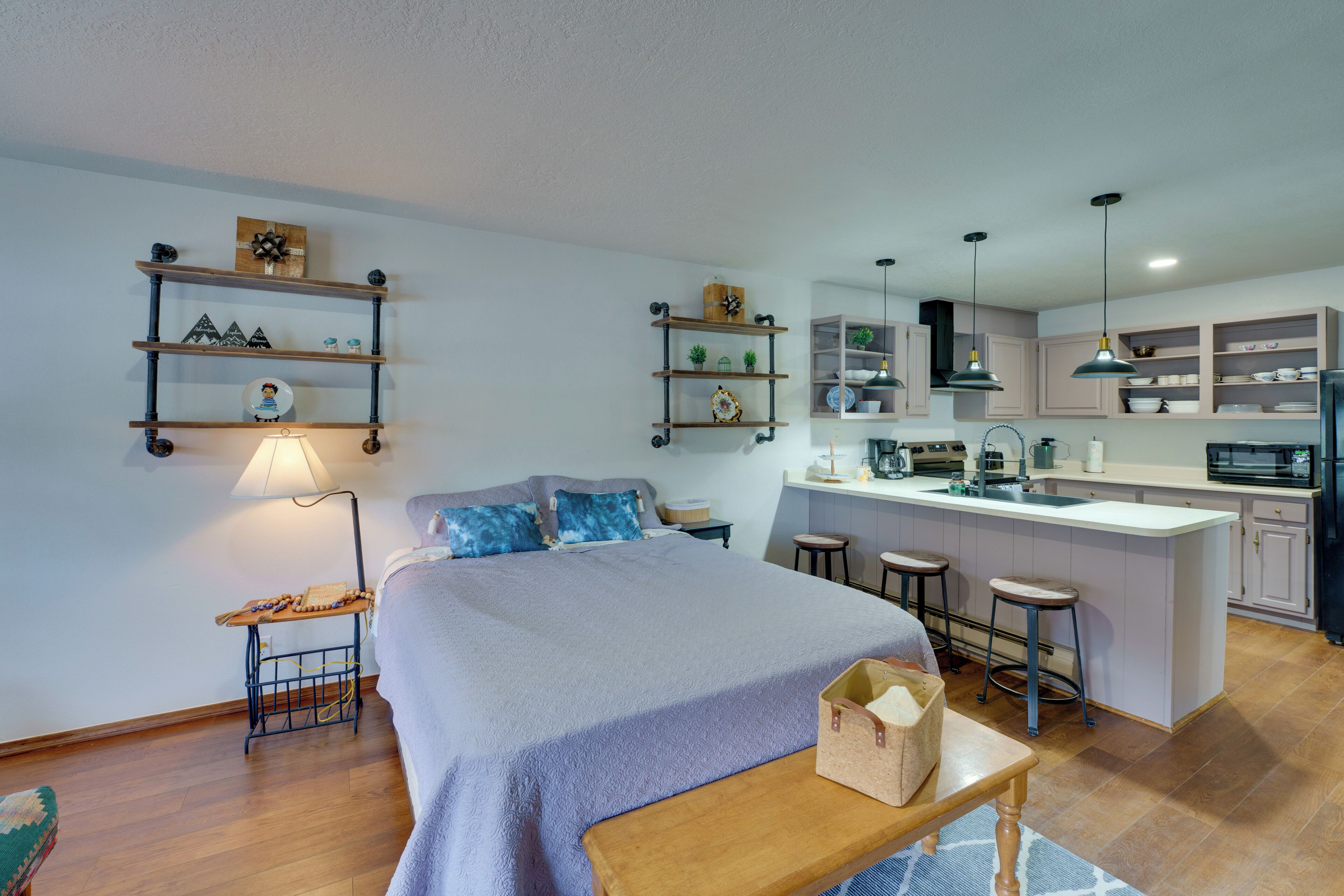 Een moderne slaapkamer met een groot bed, nachtkastjes en een klein bureau. Grenzend daar aan een compacte keuken met een centraal eiland, barkrukken en een spoelbak.