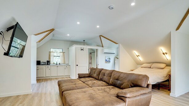 Un dormitorio tipo loft moderno con un amplio sofá de cuero marrón, una cama con manta color beige y una cocina al fondo.