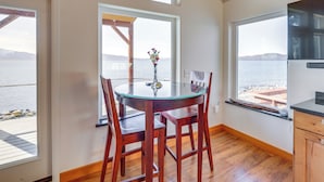 Un comedor con mesa de vidrio y sillas de madera, un jarrón con flores y vista al agua a través de amplios ventanales.