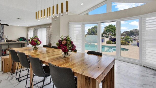 A spacious dining area with a wooden table and chairs, a vase of flowers, and a view of a swimming pool through large windows. - Waterfront Oasis – 6BR Home w/ Pool, Spa & kayaks (Largo)