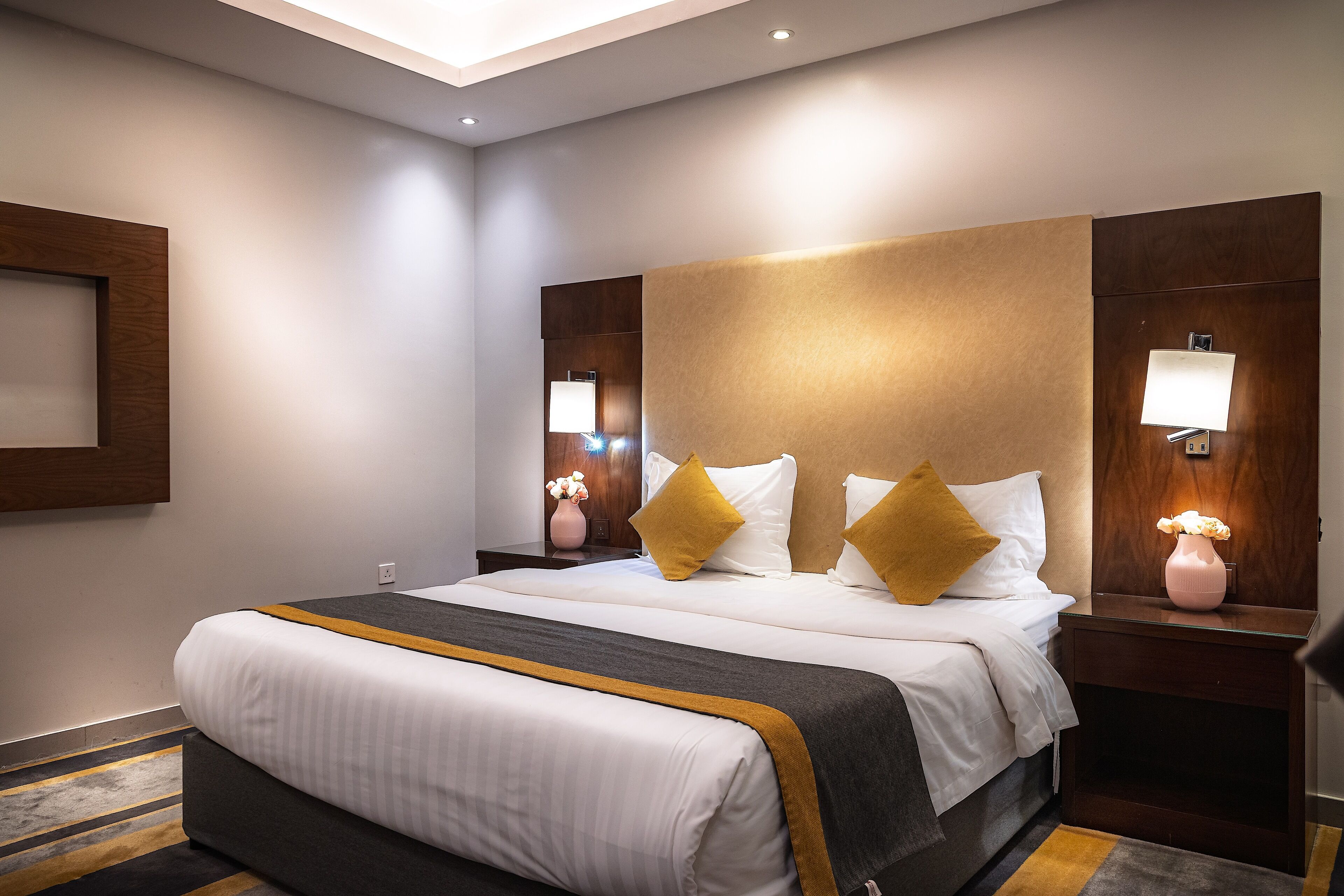 Une chambre d’hôtel avec un grand lit, deux tables de chevet avec des lampes, une tête de lit en bois et un petit vase de fleurs posé sur la table de chevet.