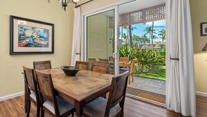 A dining area with a wooden table and chairs, a framed painting on the wall, and a view of a tropical landscape through large glass doors. - Summer Deal Waikoloa Townhome with Resort Pools Hot Tub and A-Bay (Waikoloa)