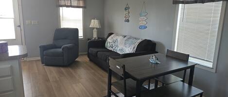 A living room with a dining table, a sofa, a chair, a ceiling fan, and a window with blinds.