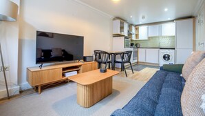 A modern apartment with a flat-screen TV, a wooden coffee table, a sofa, a kitchen area, and a washing machine. - Kensington & Earls Court 1 bed Apartment - 4 Guest (London)