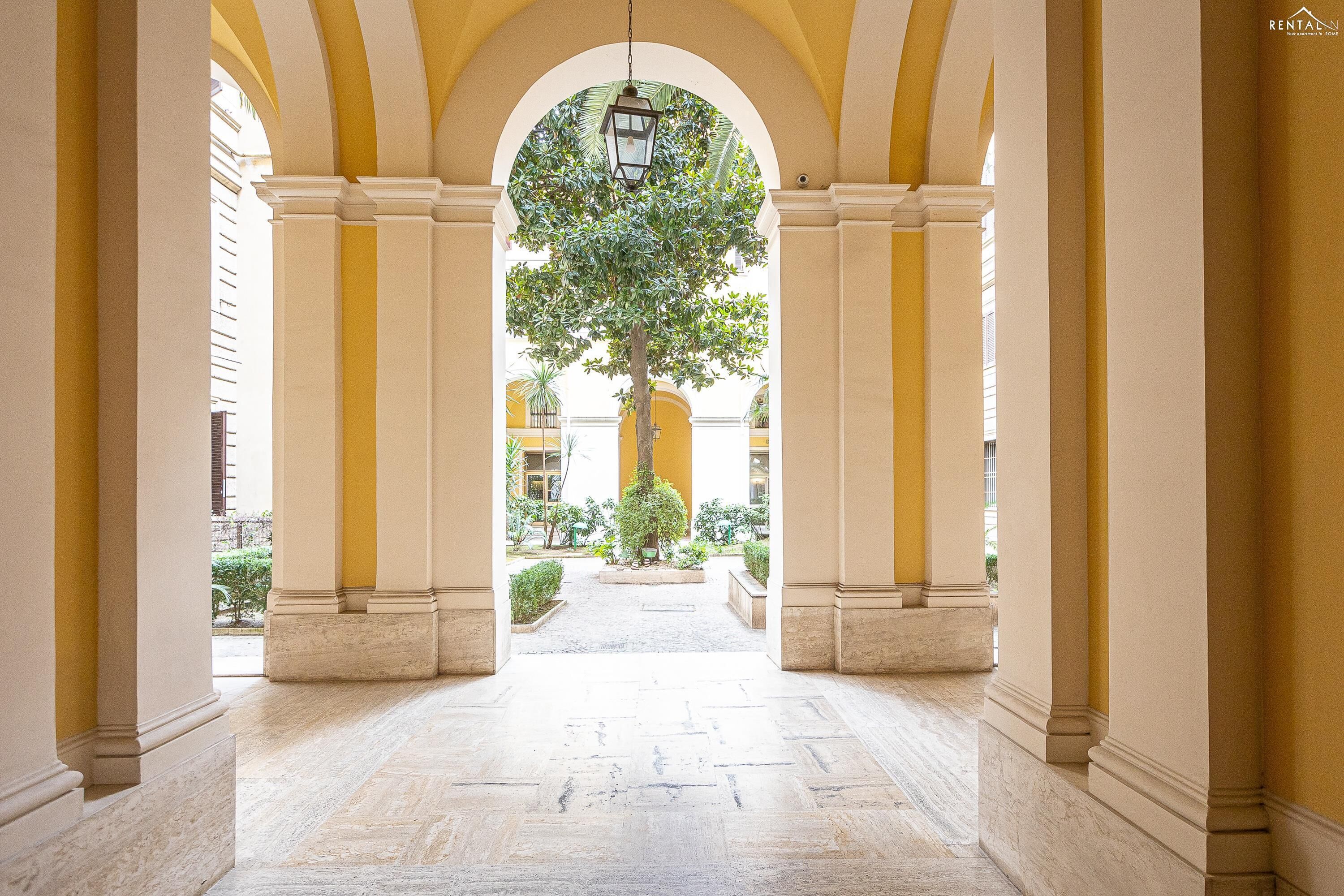 A corridor with marble flooring and yellow walls, leading to a central tree and a view of a courtyard.