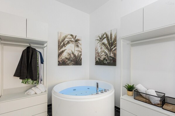 A modern bathroom with a freestanding bathtub, white cabinetry, and two wall-mounted paintings of tropical foliage.
