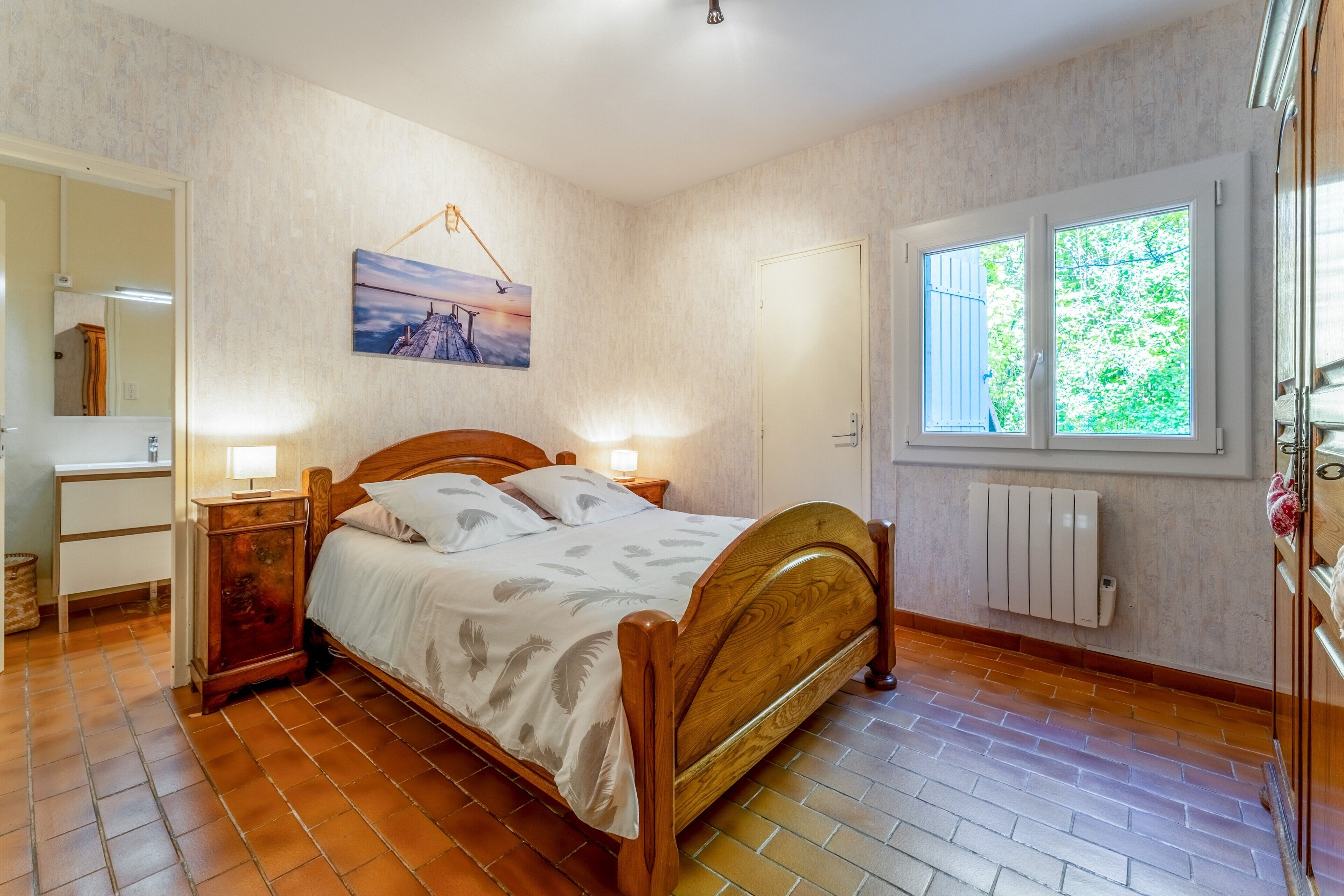 Et soveværelse med en træseng, et natbord med lampe, et vindue med udsigt til grønne omgivelser og en radiator.
