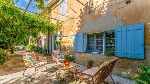 Une terrasse extérieure avec des meubles en osier, une petite table et une fenêtre ornée de volets bleus.
