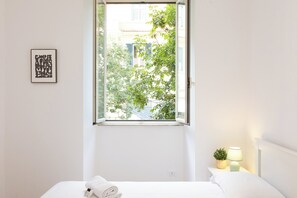 A bedroom with a large window, a bedside lamp, a small plant, and a framed artwork on the wall. - Rental in Rome Faleria Apartment (Rome)