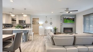 Eine moderne Küche mit weißen Schränken, einer zentralen Kochinsel und Pendelleuchten. Es gibt einen Essbereich mit Tisch und Stühlen sowie ein Wohnzimmer mit einer großen Couch und einem Kamin.