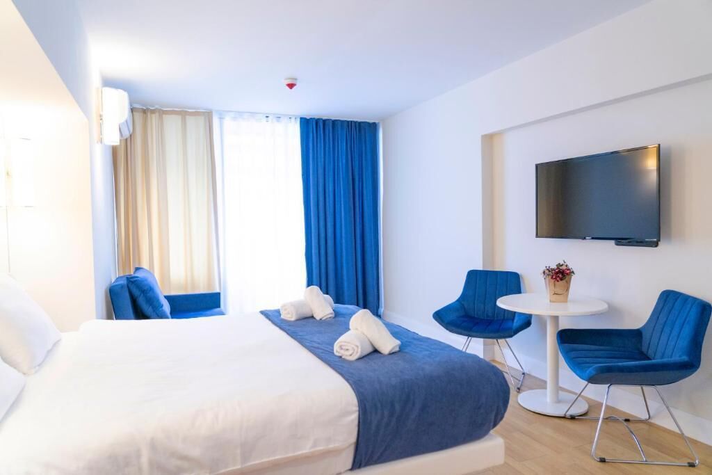 A hotel room with a bed, two blue chairs, a small white table, and a TV on the wall.