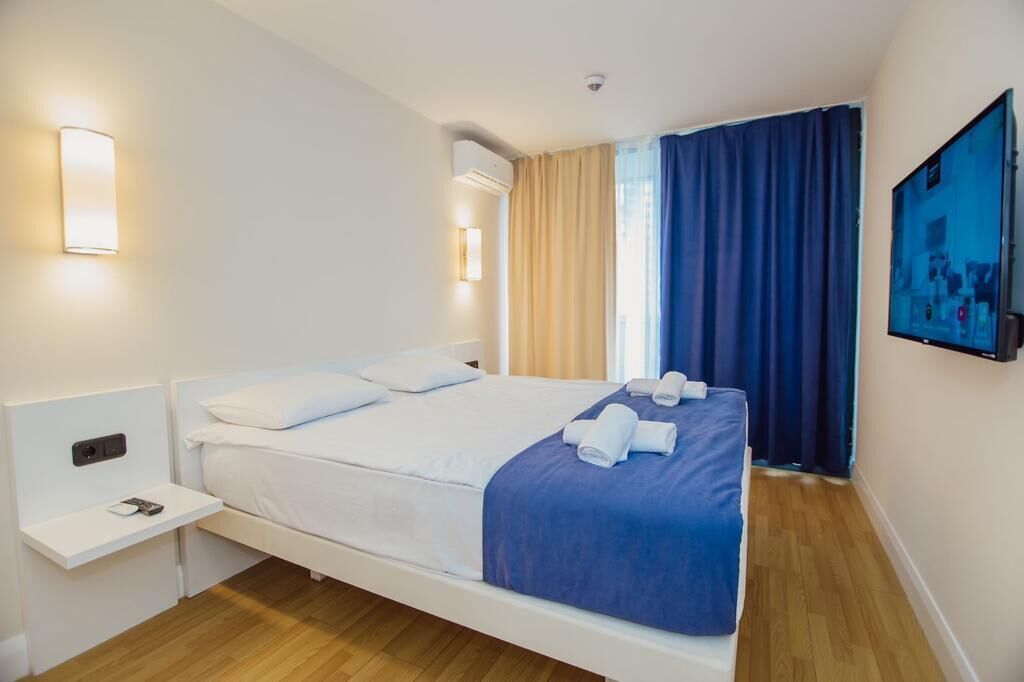 A hotel room with a bed, a bedside table with a lamp, a wall-mounted TV, and a window with curtains.