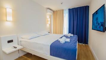 A hotel room with a bed, a bedside table with a lamp, a wall-mounted TV, and a window with curtains.