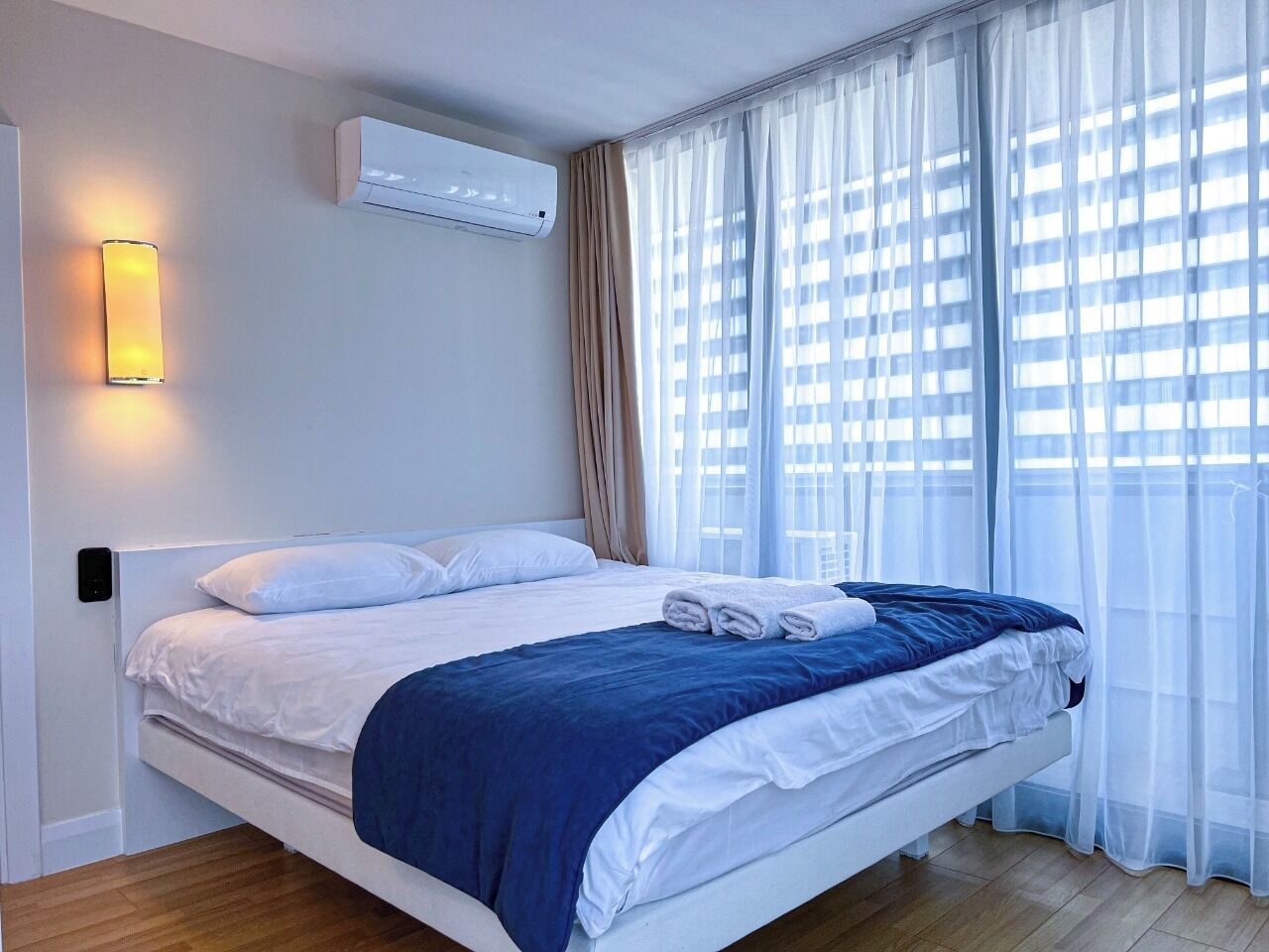 A modern hotel room with a bed, air conditioning unit, and a view of buildings through sheer curtains.