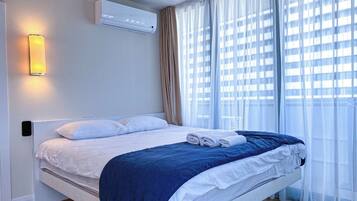 A modern hotel room with a bed, air conditioning unit, and a view of buildings through sheer curtains.
