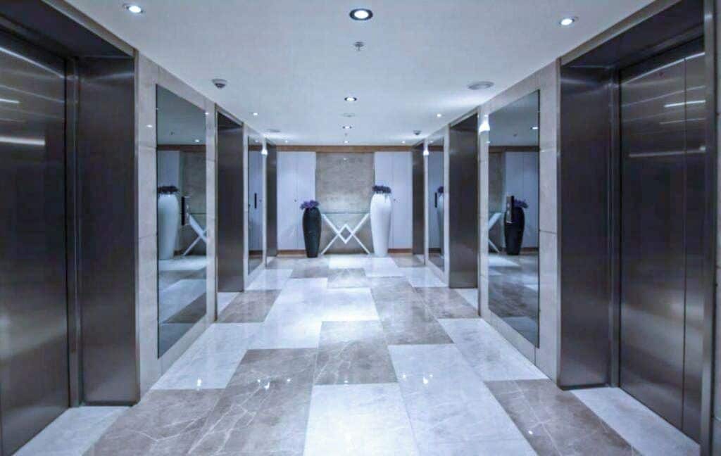 A modern hallway with marble flooring, glass walls, and elevators.