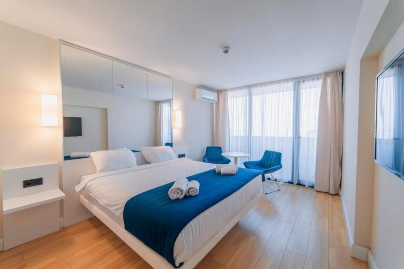 A modern hotel room with a large bed, a sitting area with a table and chairs, a large mirror, and a balcony with curtains.