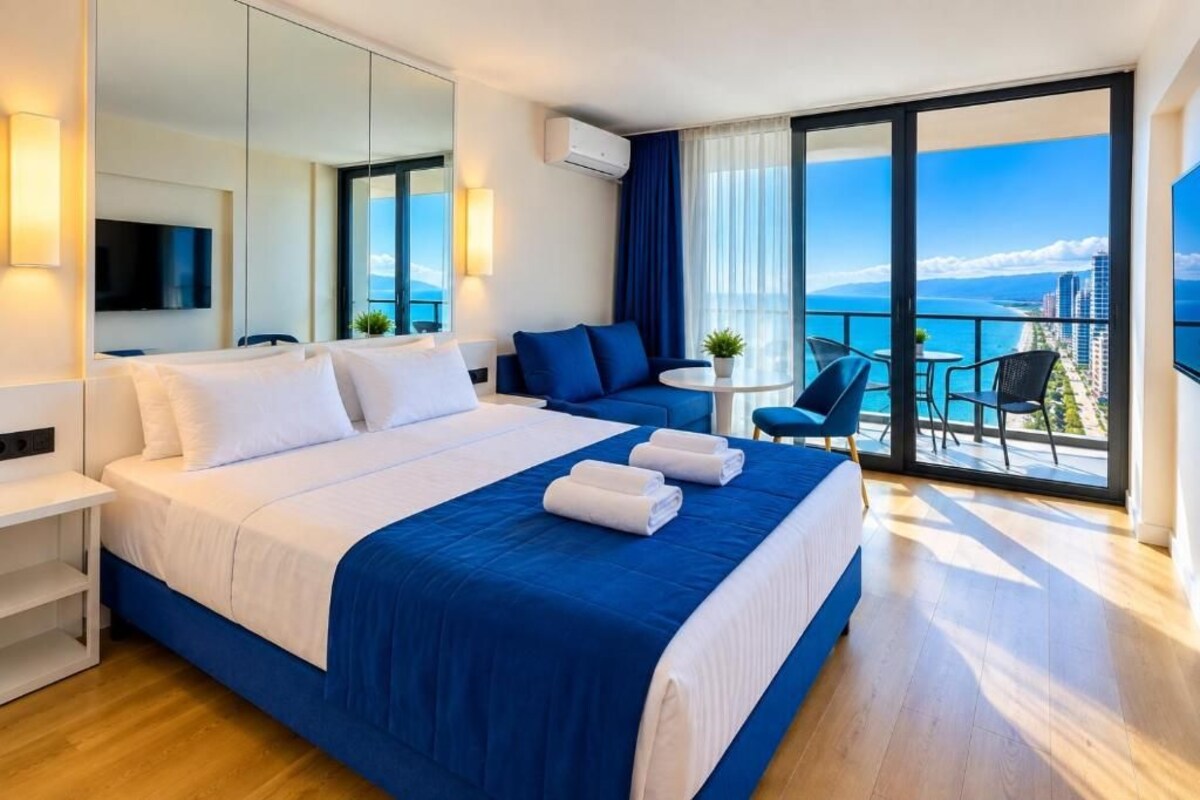 A hotel room with a large bed, a balcony with a view of the sea, a small table with chairs, and a TV mounted on the wall.