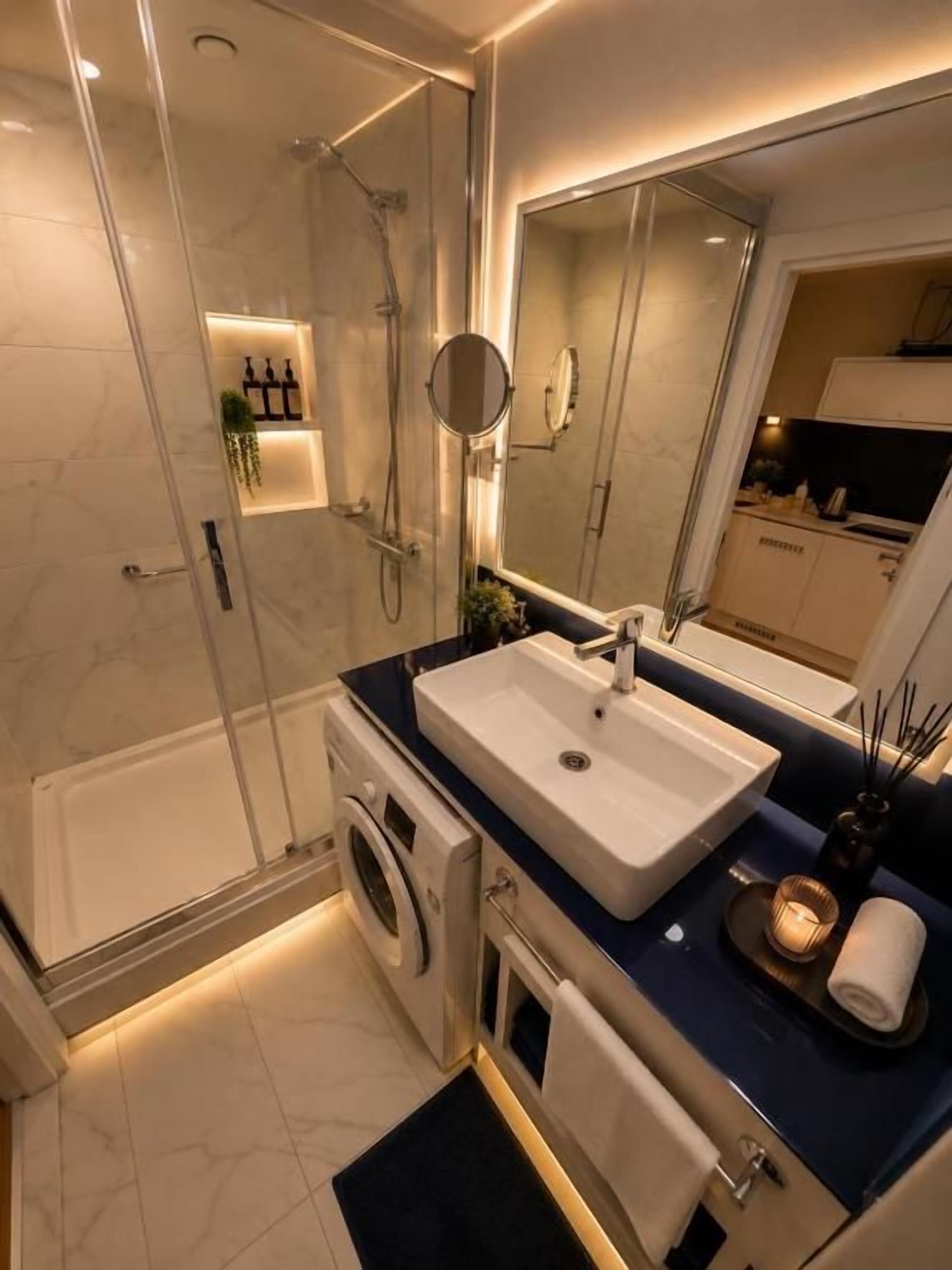 A modern bathroom with a glass shower enclosure, a built-in washing machine, a sink with a mirror, and a towel rack.