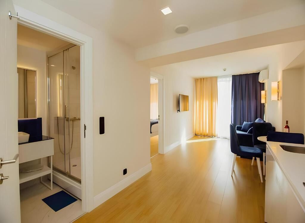 A modern hotel room with a glass shower, a dining area with a table and chairs, and a living area with a sofa and a TV.
